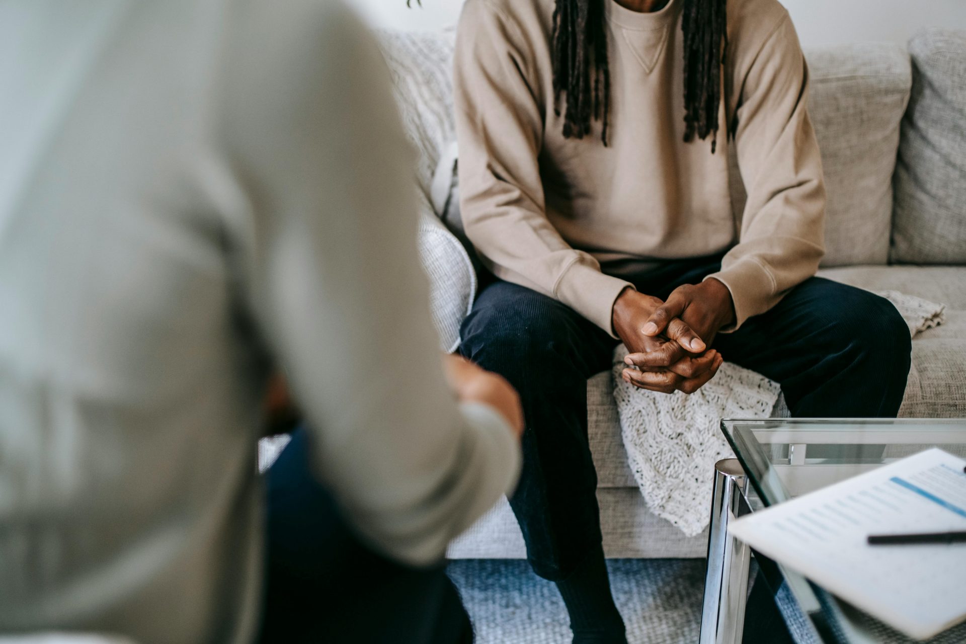 Patient afro-américain de sexe masculin, non identifiable, les mains jointes, assis sur un canapé près d'un thérapeute anonyme flou lors d'une consultation en cabinet de psychothérapie.